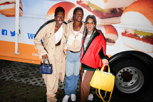 Telfar Clemens stands with two women, they are all smiling and laughing, full of joy and carring a small navy and large yellow Telfar bags in front of a sandwich food truck.